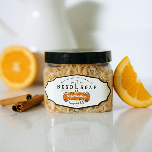 The Tangerine Spice Milk Bath jar is displayed on a white surface with orange slices and cinnamon sticks, emphasizing its skin rejuvenating benefits from tangerine oil.