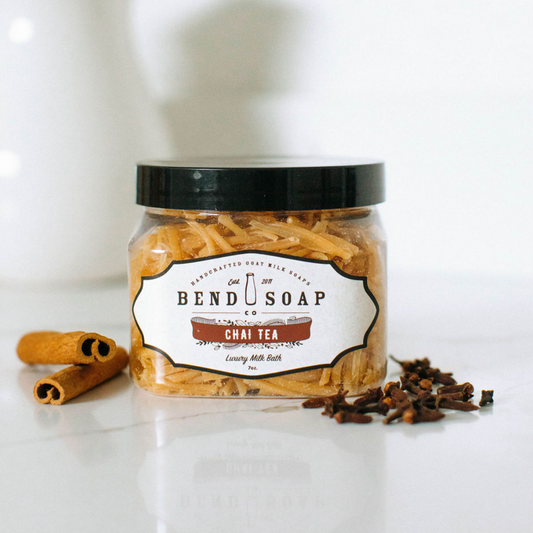 A clear jar labeled "Chai Tea Milk Bath" filled with tan soap shreds sits on a white surface, bringing a cozy cinnamon chai aroma to your self-care routine, with two cinnamon sticks and cloves beside it.