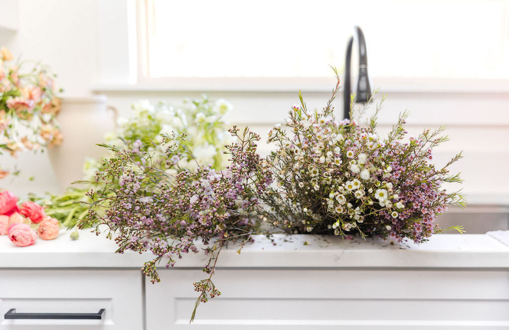 A bouquet of small white and light pink flowers with green stems rests on a white kitchen counter in front of a sink, with more flowers and greenery in the blurred background.