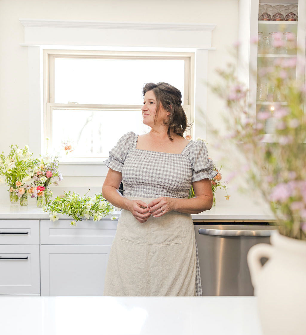 A woman in a checkered dress and apron stands in a bright kitchen with white cabinets, surrounded by fresh flowers on the counter, looking out a window.