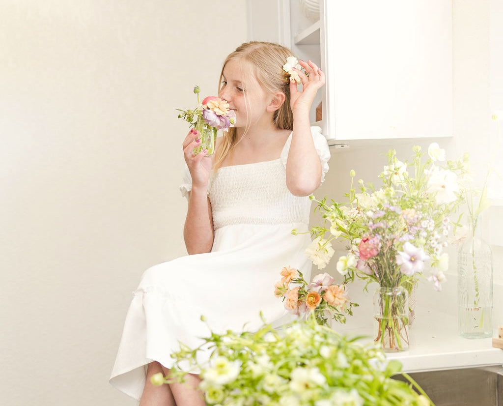 A young girl in a white dress sits on a counter, holding and smelling a bouquet of flowers. More flowers are arranged around her, creating a bright and fresh atmosphere.