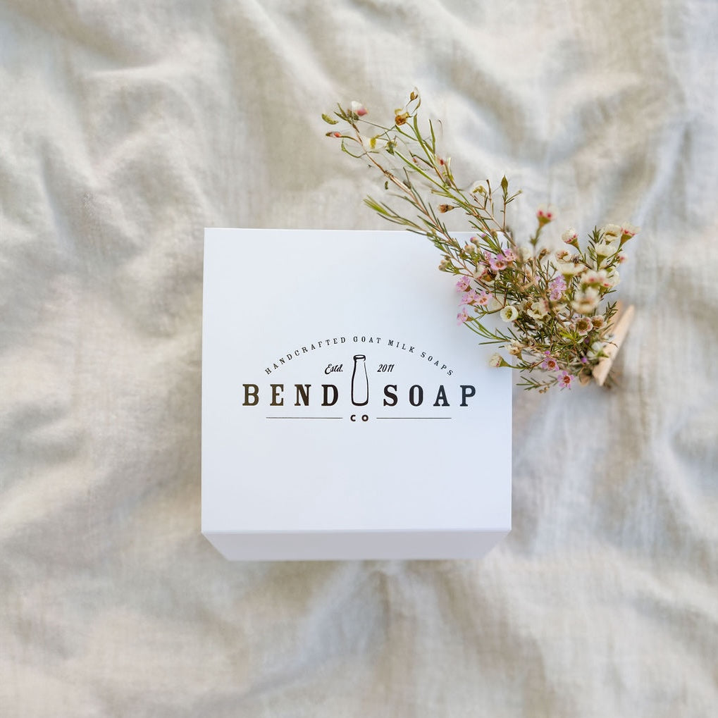 A white Bend Soap Co. box sits on a light-colored fabric, with a small sprig of delicate pink and white flowers resting on top.