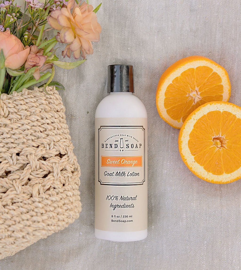 A bottle of Bend Soap Co. Sweet Orange Goat Milk Lotion sits on a textured surface beside two orange slices and a woven basket with peach and pink flowers.