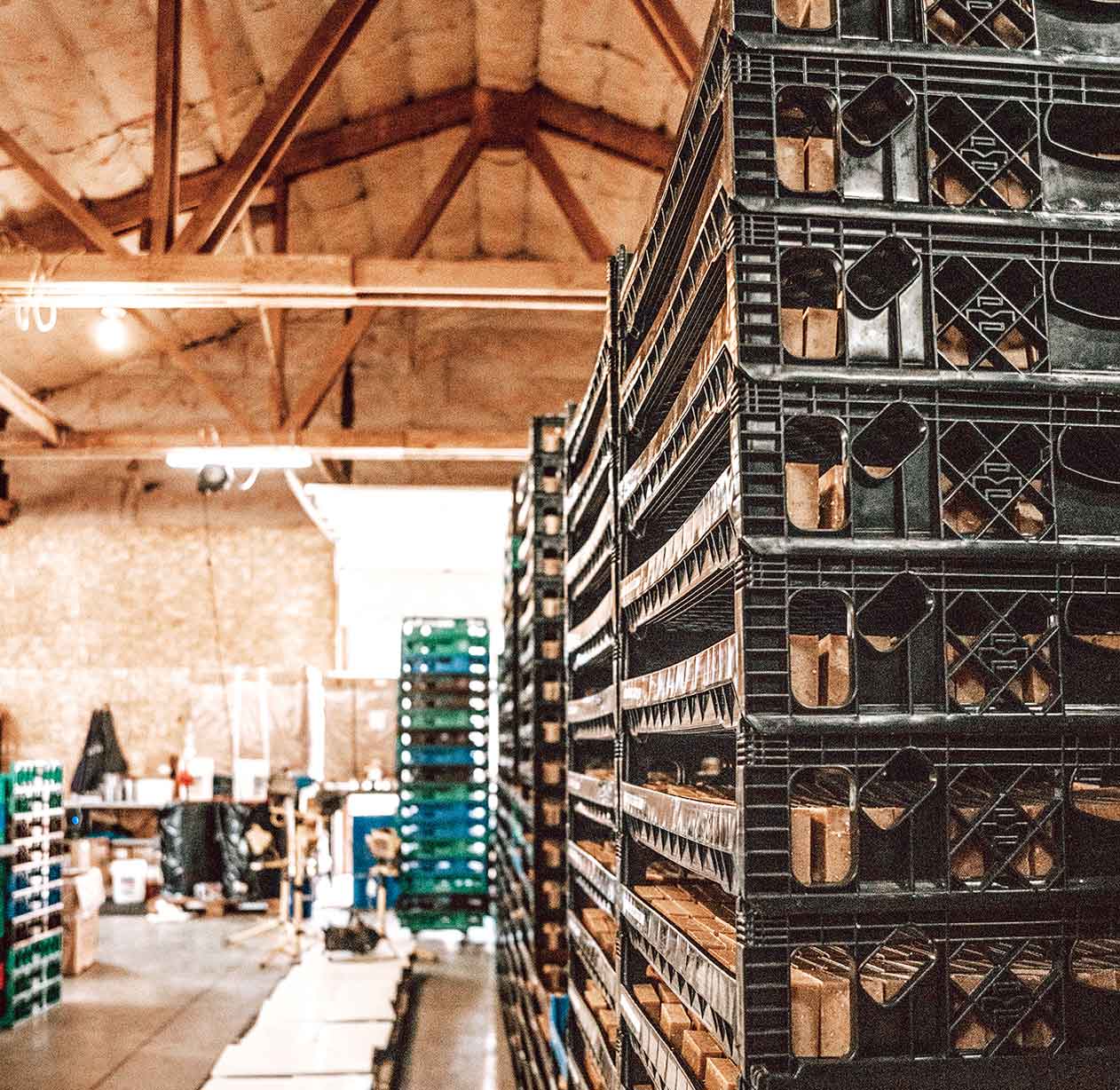 Stacks of black plastic crates are arranged in a warehouse with wooden beams on the ceiling and various storage items and equipment visible in the background.