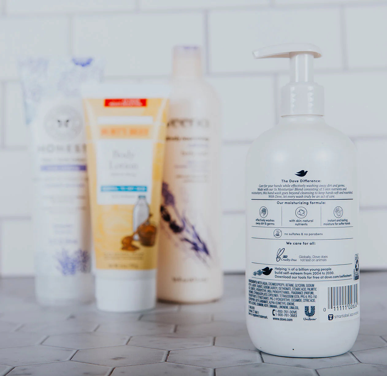Four lotion bottles are arranged on a tiled surface, with one white pump bottle in sharp focus showing detailed text, while three other bottles in various colors and shapes are blurred in the background.