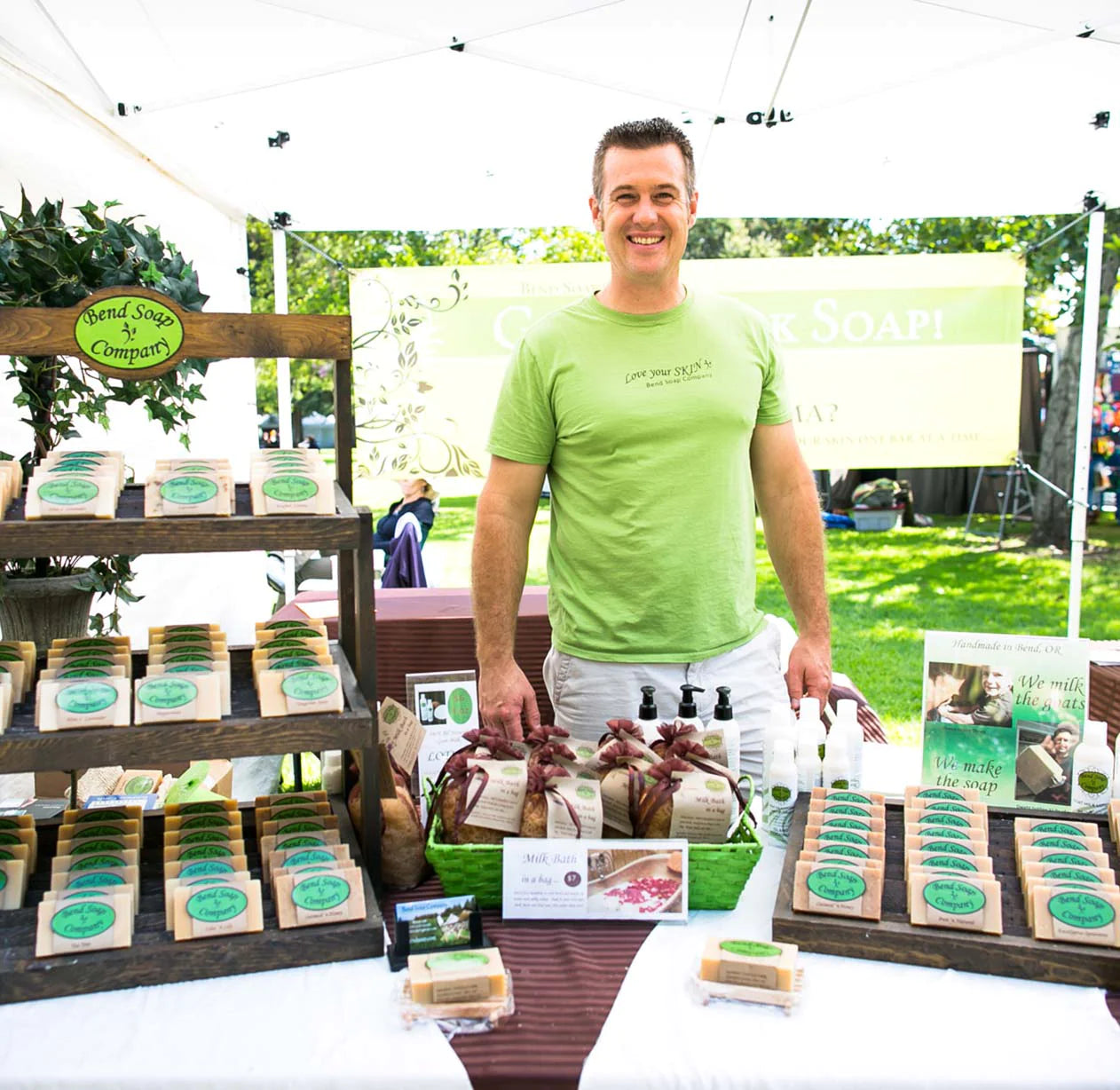A smiling man in a light green shirt stands behind a farmer’s market booth displaying handmade soaps arranged on wooden racks, with a Bend Soap Company sign and a sunny outdoor setting in the background.