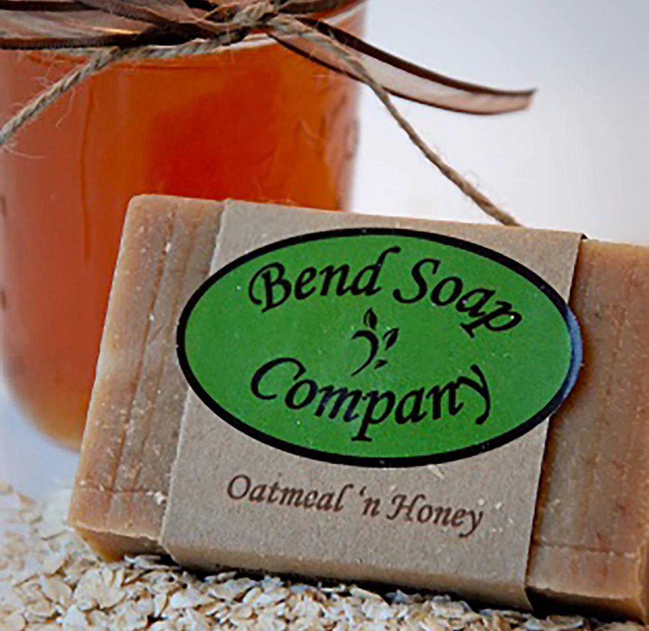 A bar of Bend Soap Company oatmeal 'n honey soap sits on oats, with a jar of honey tied with string in the background.
