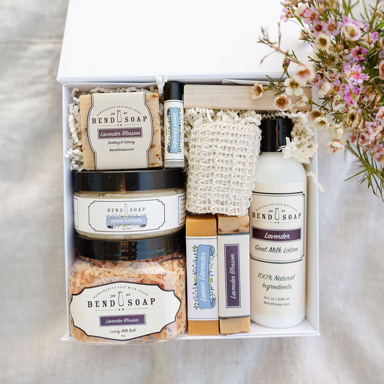 The Blooming Blossom Box includes Bend Soap Co. lotion, soap bars, bath soak, lip balm, a loofah, and a wooden soap dish—beautifully arranged with flowers—for the perfect spa day gift at home.