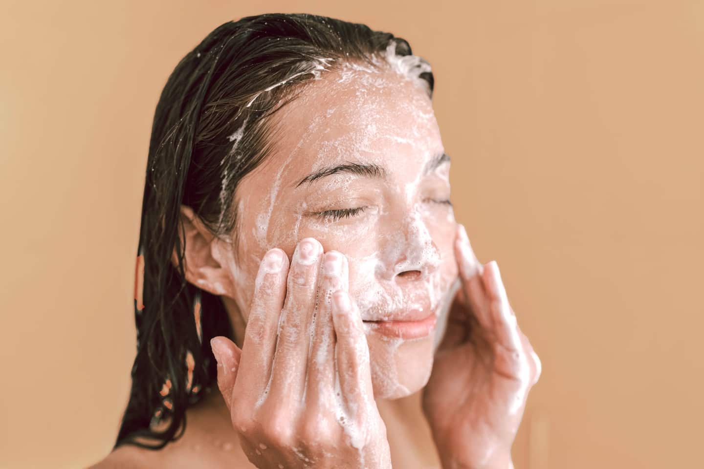 A person with wet hair gently massages foaming cleanser onto their face with closed eyes, standing against a plain beige background.