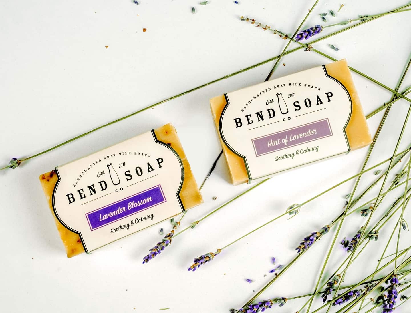 Two bars of Bend Soap Co. goat milk soap, labeled "Lavender Blossom" and "Hint of Lavender," lie on a white surface surrounded by scattered sprigs of fresh lavender.