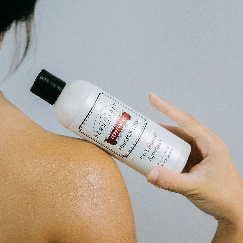 Someone holds an 8oz bottle of Peppermint Goat Milk Lotion near their bare shoulder, showcasing this moisturizer’s skin health benefits against a light gray background.