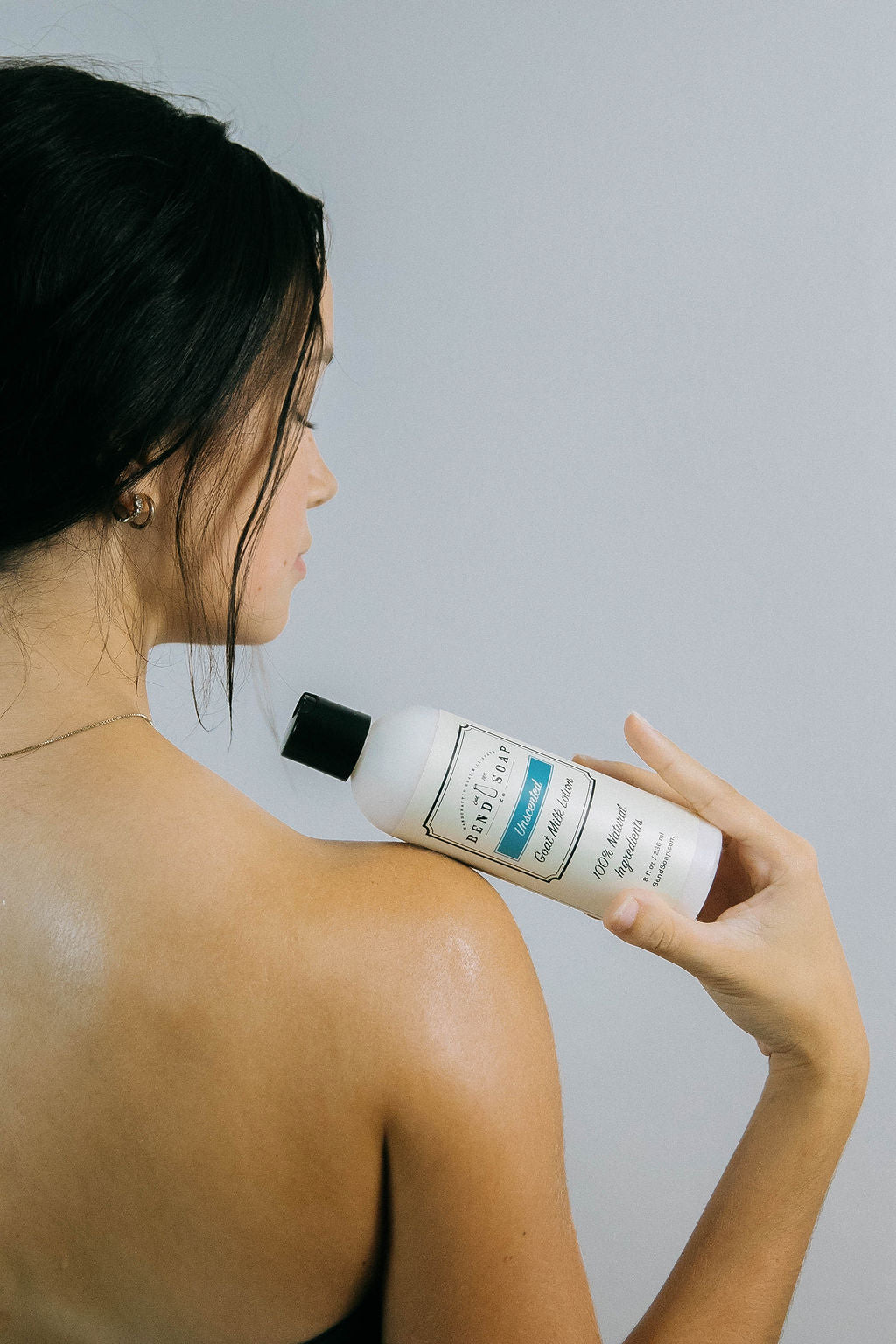 A woman with dark hair holds a white bottle from the Best for Acne Bundle near her bare shoulder, looking away against a light background and showcasing her clear, acne-free skin.