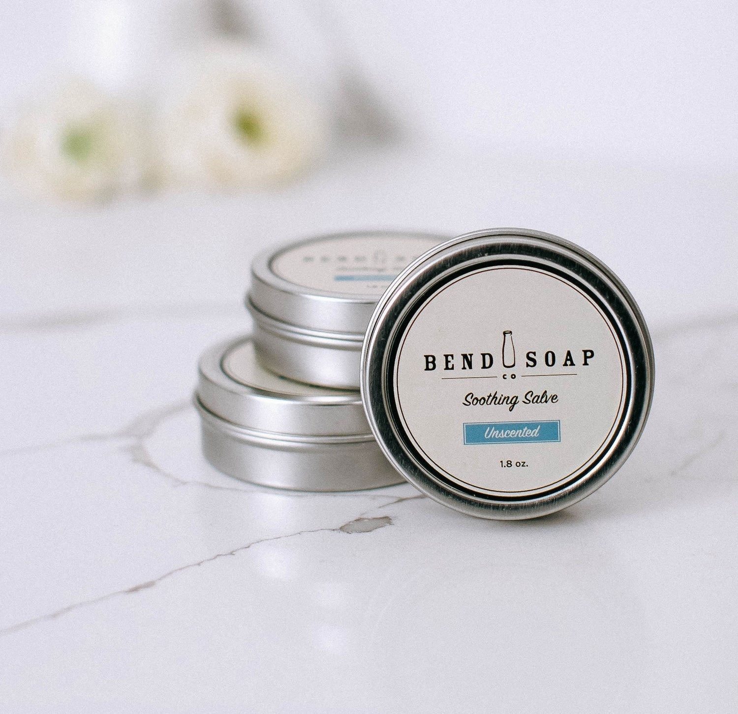Three silver tins of Bend Soap Soothing Salve, labeled “Unscented,” are stacked on a white marble surface with a soft, blurred floral background. The front tin’s label is clearly visible.