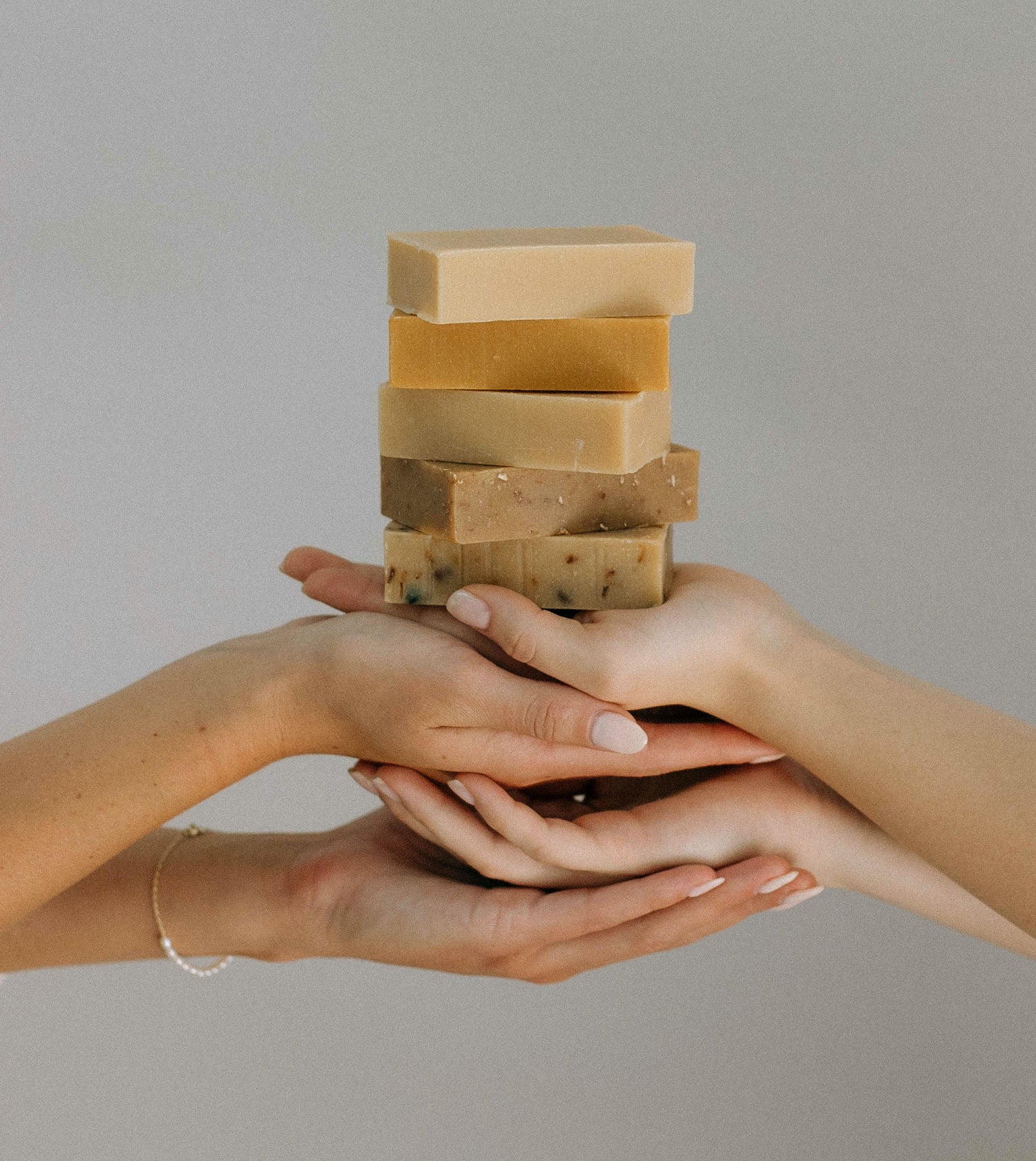 Several hands gently hold a stack of natural, handmade soap bars in various earthy colors against a plain, light background.