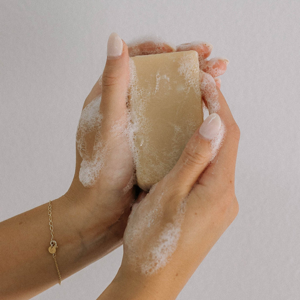 A person with neatly manicured nails holds a lathered bar from the 12 Pack of Imperfect Bars - Honey Grapefruit Goat Milk Soap, against a plain light background, wearing a delicate gold bracelet to highlight the soap’s moisturizing qualities.