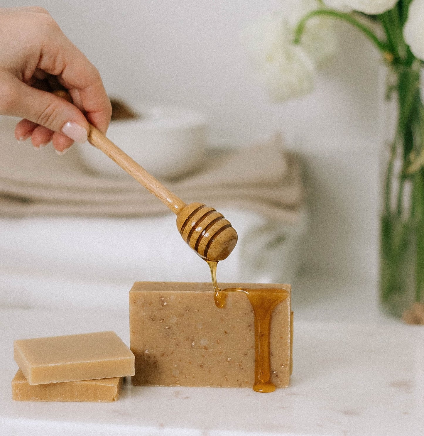 A hand drizzles honey over a bar from the 12 Pack of Imperfect Bars - Oatmeal & Honey Goat Milk Soap, with two more bars nearby on a white surface and soft white flowers in the background. Perfect for sensitive skin.