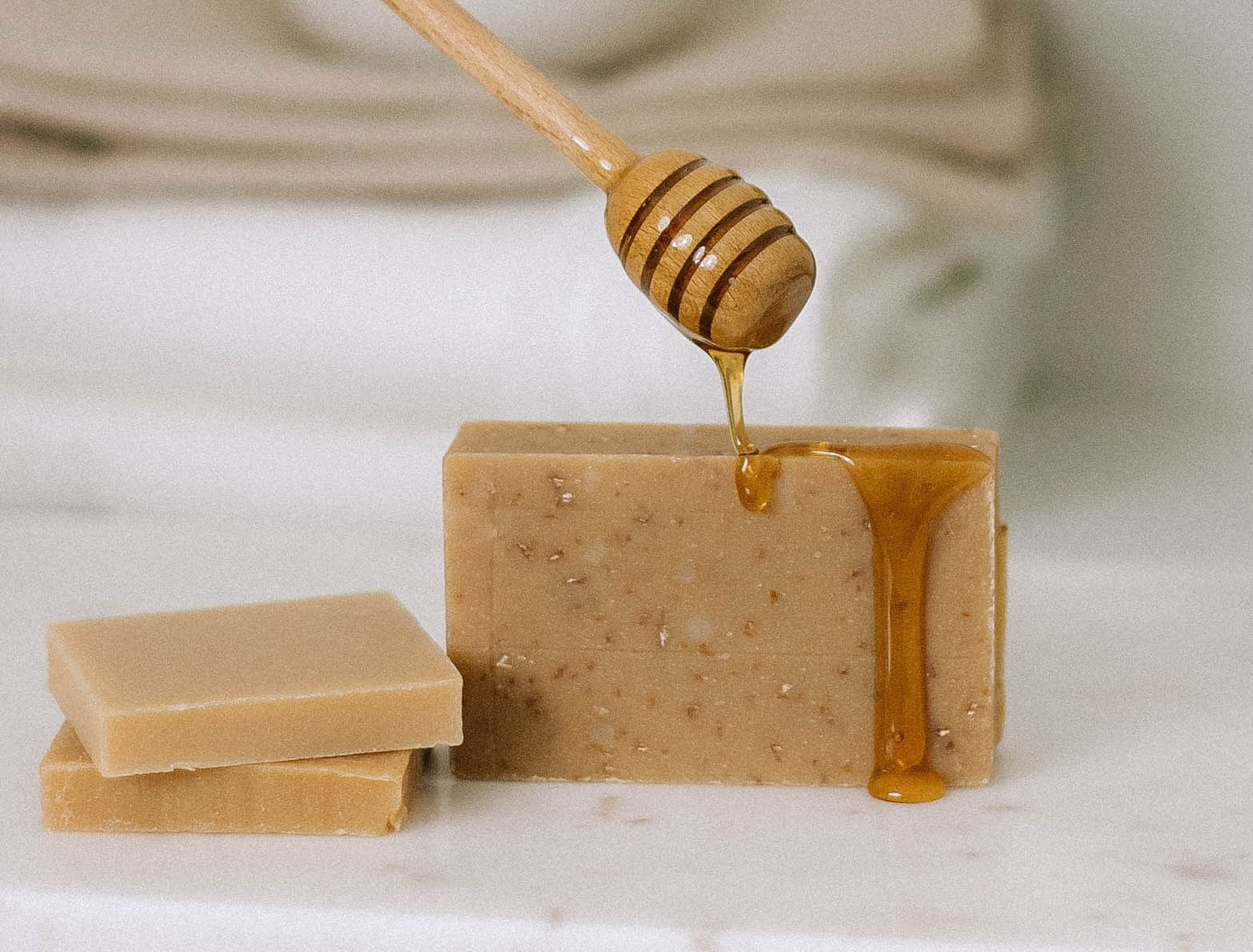 A honey dipper drizzles honey over a bar of natural soap, with two other soap bars stacked nearby on a white surface. Soft, neutral backgrounds create a calming atmosphere.
