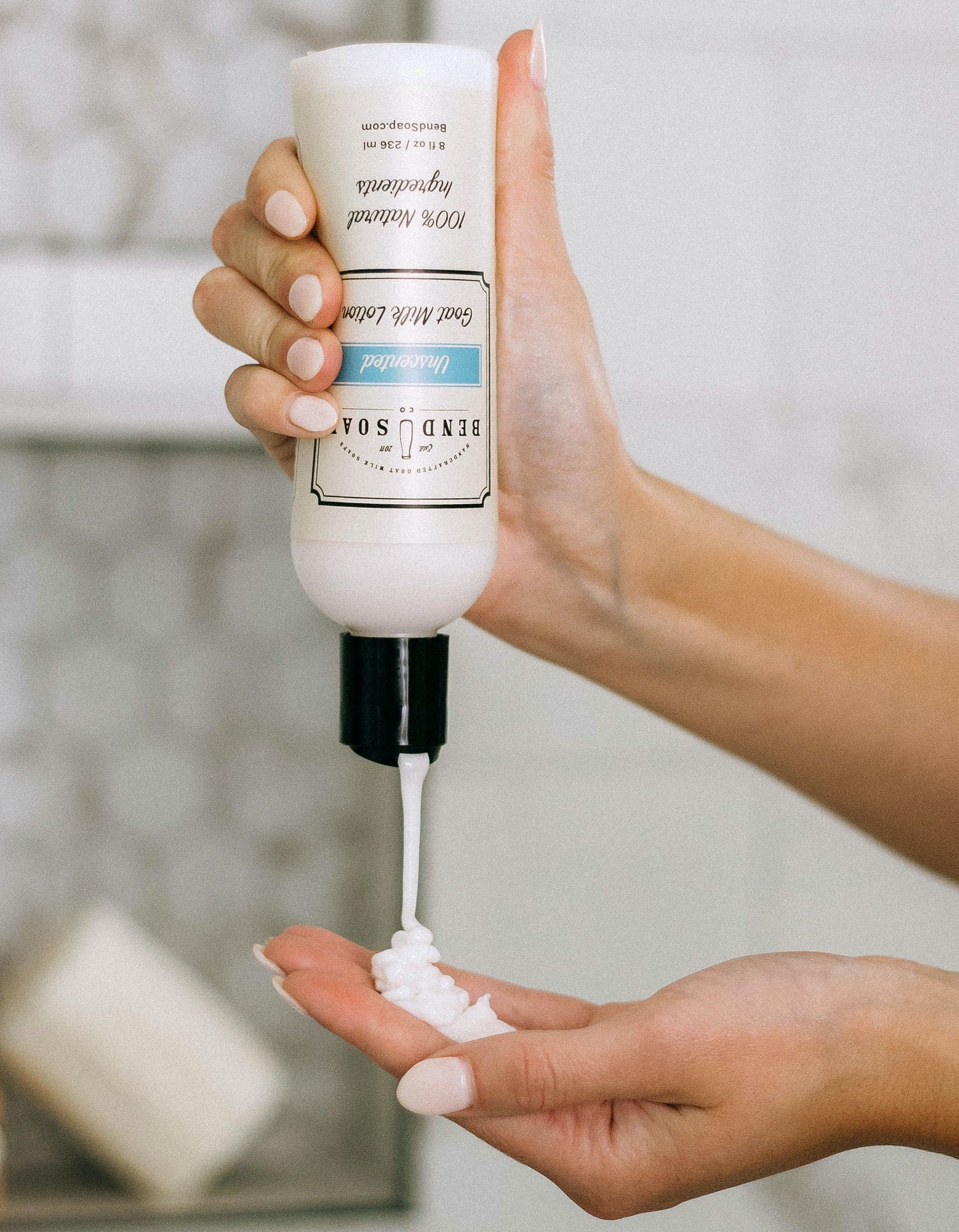 A person dispenses white lotion from a tube labeled "Bend Soap Co." into their hand, with a blurred soap bar in the background.