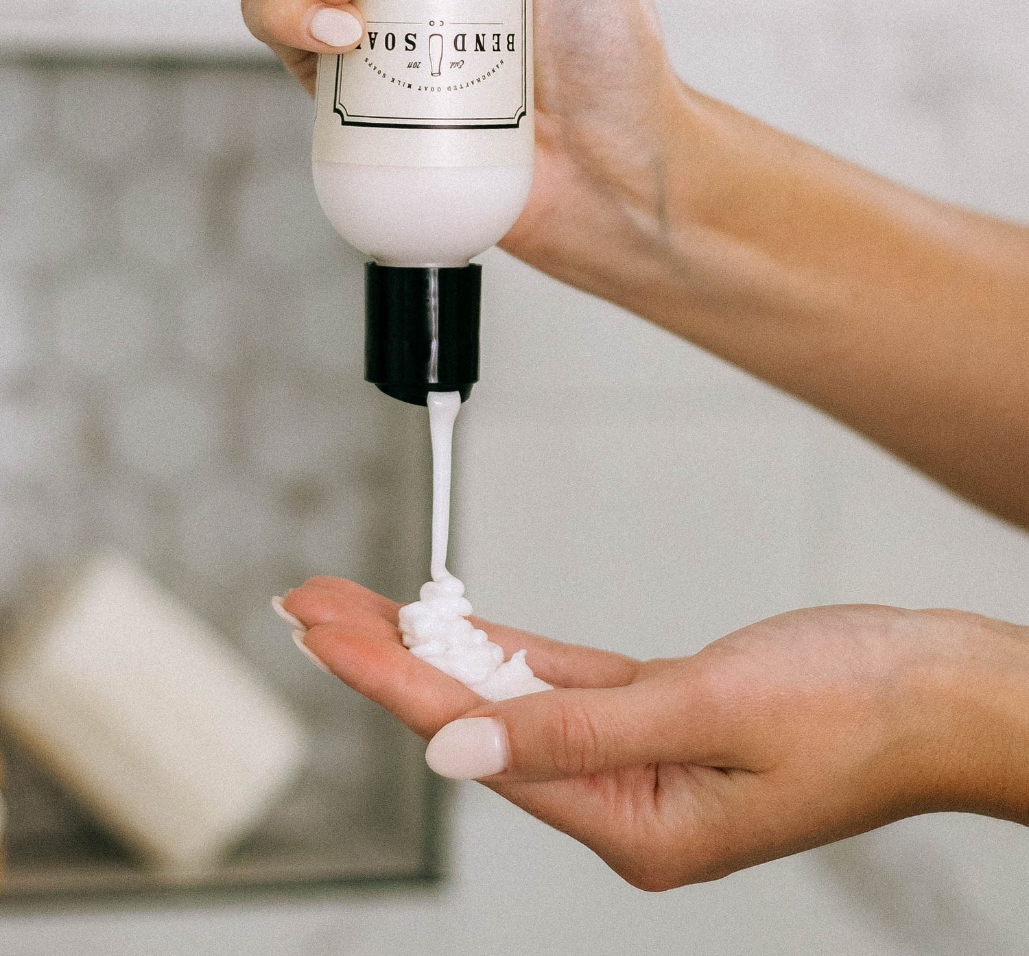 A person dispenses white lotion from a bottle onto their palm in a bathroom, with a soap dish visible in the background.