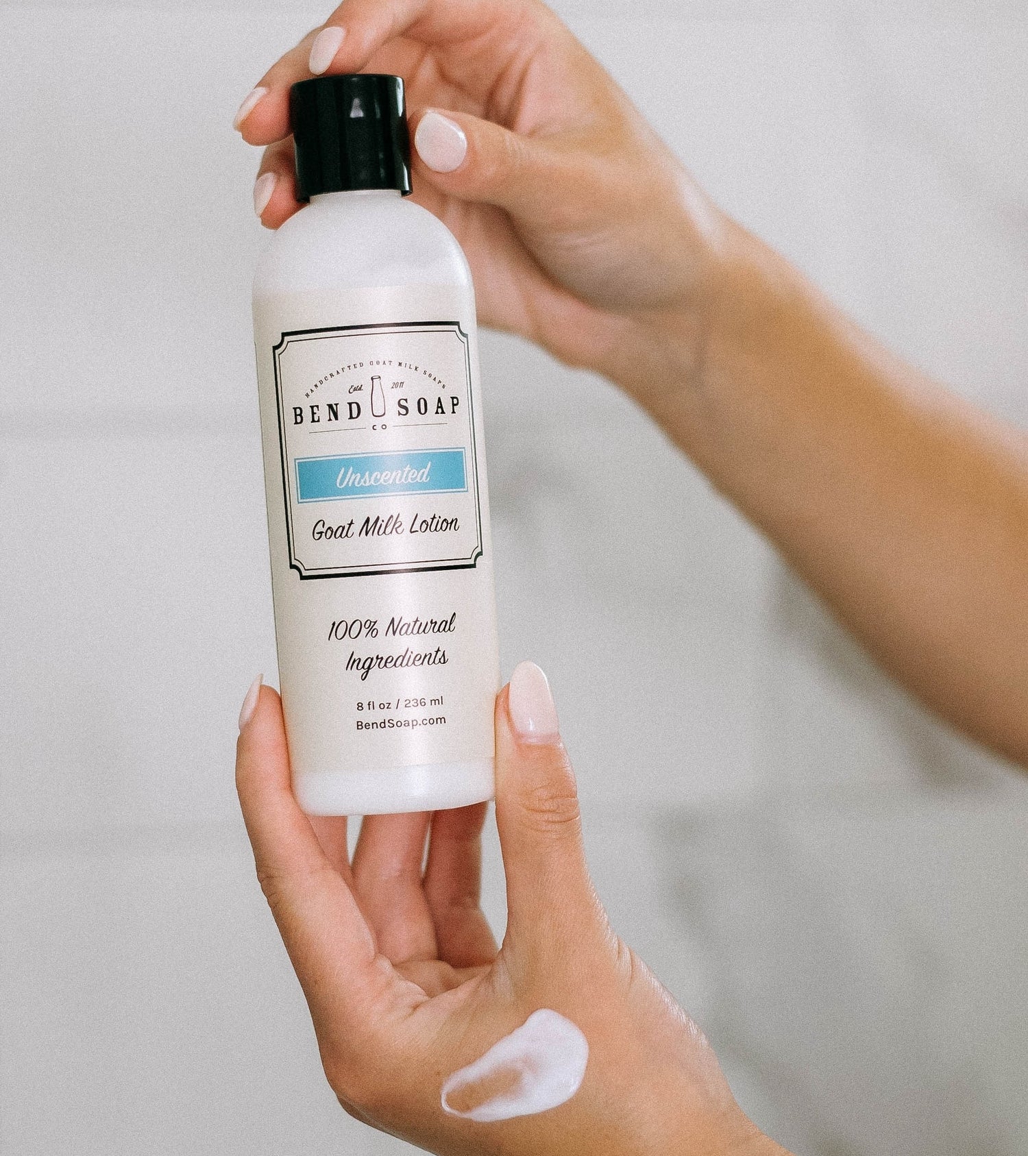 A person holds a bottle of Bend Soap Company Unscented Goat Milk Lotion in one hand, with a dab of lotion on the back of their other hand. The bottle label indicates it contains 100% natural ingredients.