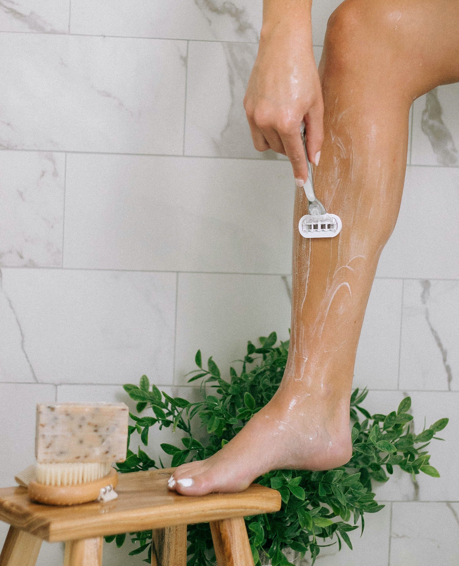 A person shaves their lower leg with a razor, leg propped on a wooden stool. Soap, a brush, and a leafy plant are nearby, with a tiled wall in the background.
