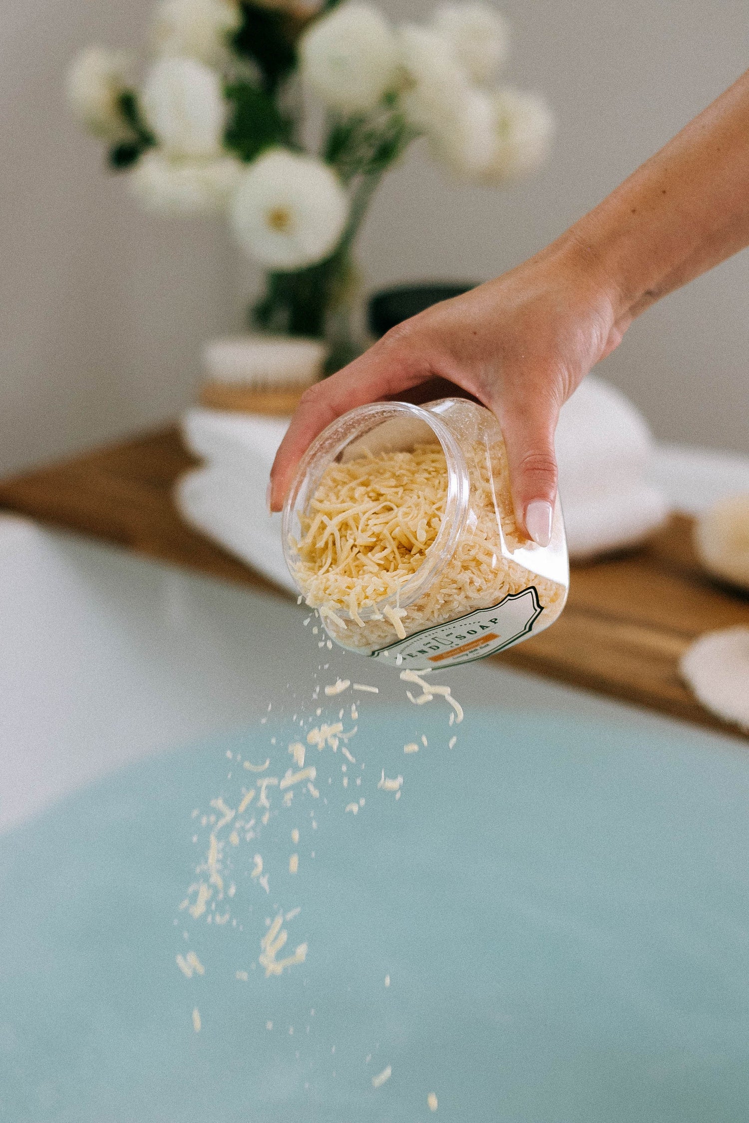 A hand pours shredded cheese from a jar into a bathtub filled with water, with flowers and a wooden tray in the background.