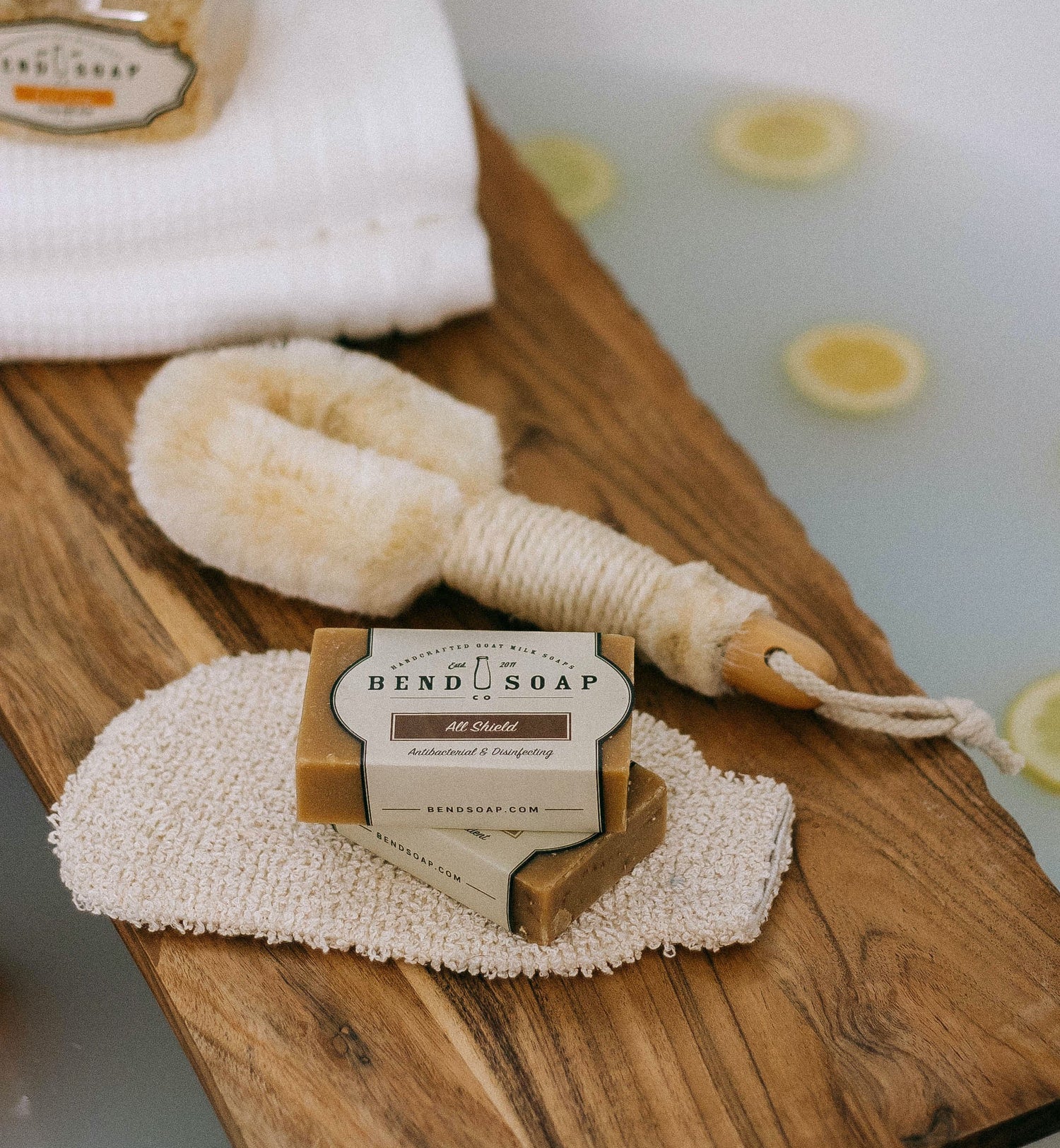 A bar of Bend Soap rests on a textured washcloth alongside a bath brush on a wooden bath tray, with a towel and lemon slices floating in bathwater in the background.