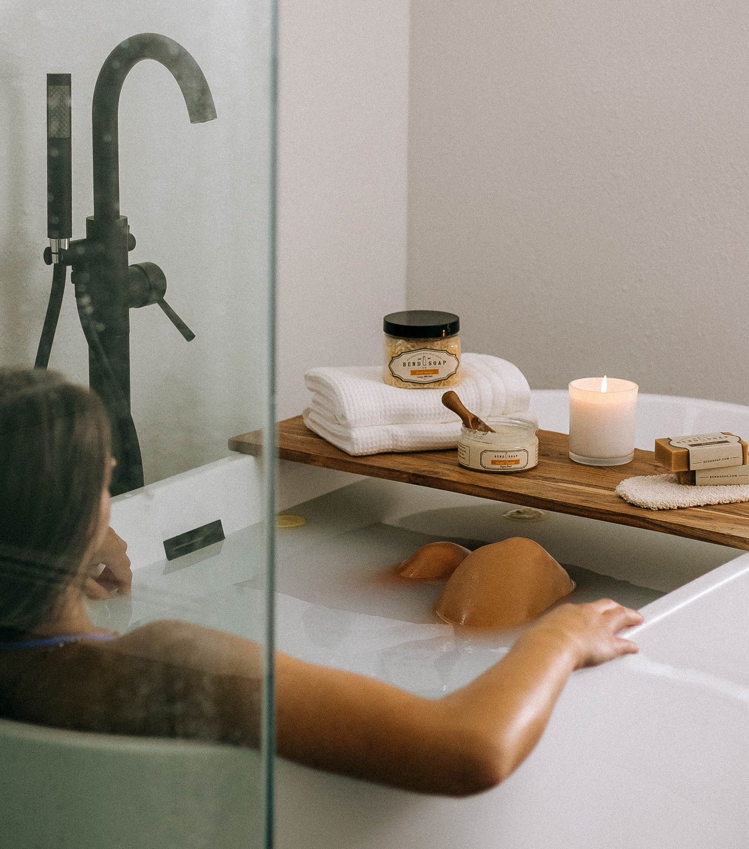 A person relaxes in a bathtub with a wooden tray holding a lit candle, skincare products, soap, a brush, and folded towels. The scene is calming and serene, suggesting a spa-like atmosphere.