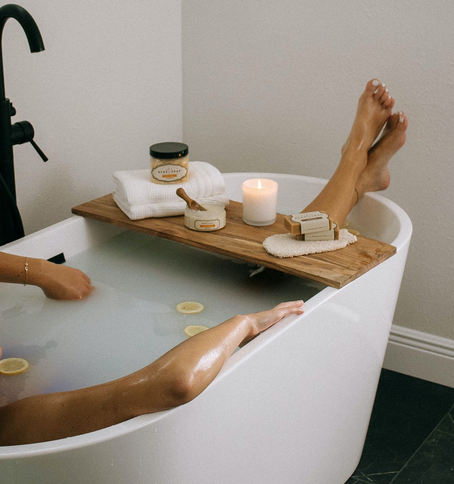 A person relaxes in a white bathtub with cloudy water and floating lemon slices; one leg rests on a wooden tray holding a candle, towel, skincare products, and a body brush.