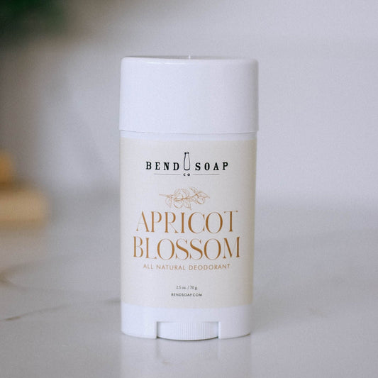 A white stick of Apricot Blossom Magnesium Deodorant (Baking Soda Free) rests on a light surface. The label has gold lettering and a simple floral design, emphasizing gentle odor protection with magnesium hydroxide.