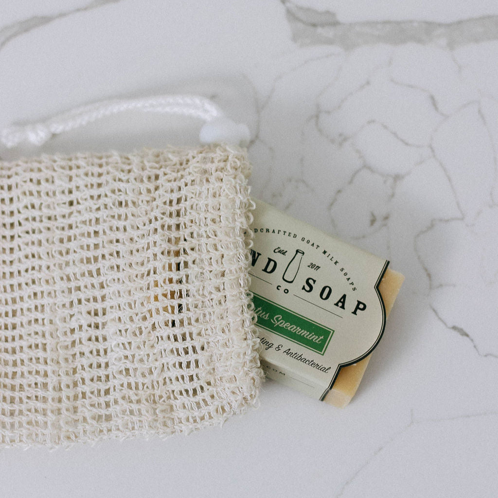 An All Natural Loofah Bag holding a bar of Hand Soap Co. Eucalyptus Spearmint soap rests on a white, cracked marble surface.