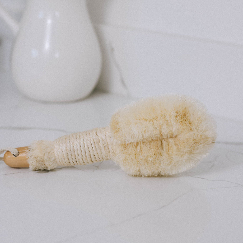 A Natural Foot Scrub Brush with a wooden handle and dense, light bristles rests on a white marble surface, ideal for exfoliating feet. A blurred white ceramic object appears in the background.