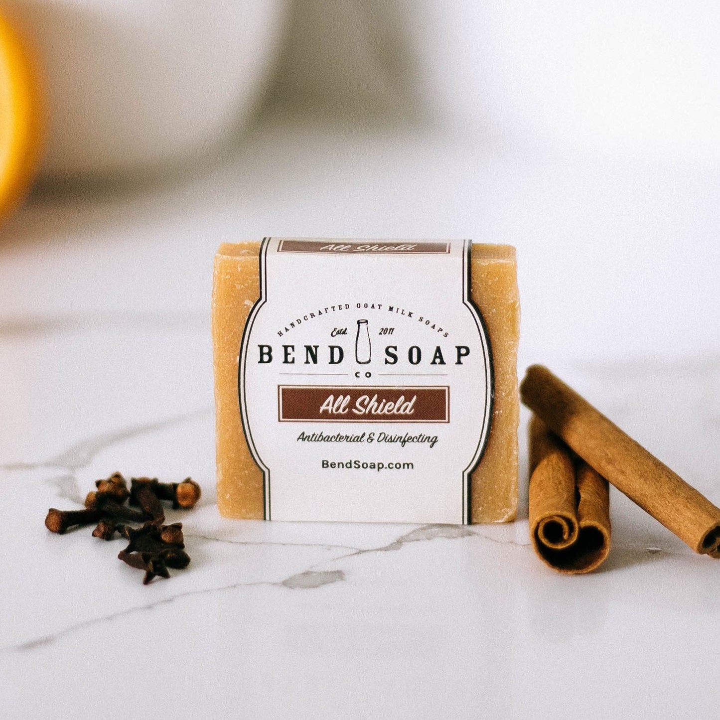 A bar of All Shield Goat Milk Soap rests on a white surface beside whole cloves and two cinnamon sticks, emphasizing its antibacterial qualities. The soap’s wrapper is white with brown detailing.