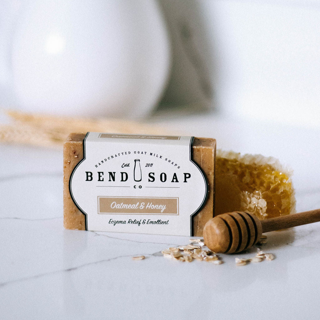 A 1 lb. batch of Oatmeal and Honey Goat Milk Soap Scrap rests on a white surface with a honey dipper, honeycomb, oats, and wheat—made with natural ingredients, ideal for sensitive skin, set against a white tiled background.