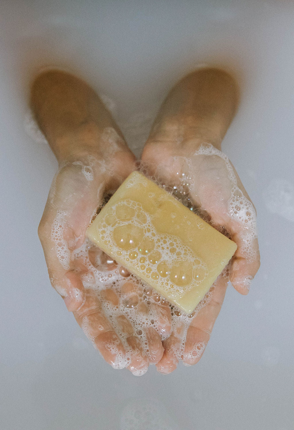 Two hands gently hold a bubbly yellow bar from the Best for Babies Bundle, submerged in soapy water with suds floating on the surface.