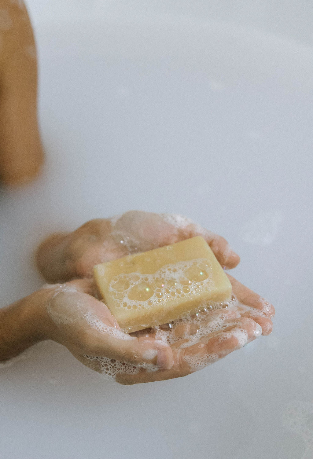 A person’s hands hold a yellow bar of Best for Acne Bundle soap with bubbles, partially submerged in milky bathwater for acne-free skin.