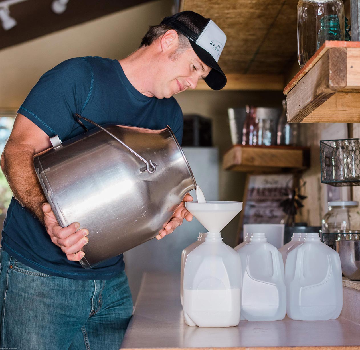 A man wearing a cap pours milk from a large metal container through a funnel into one of three plastic gallon jugs on a counter in a rustic kitchen setting.
