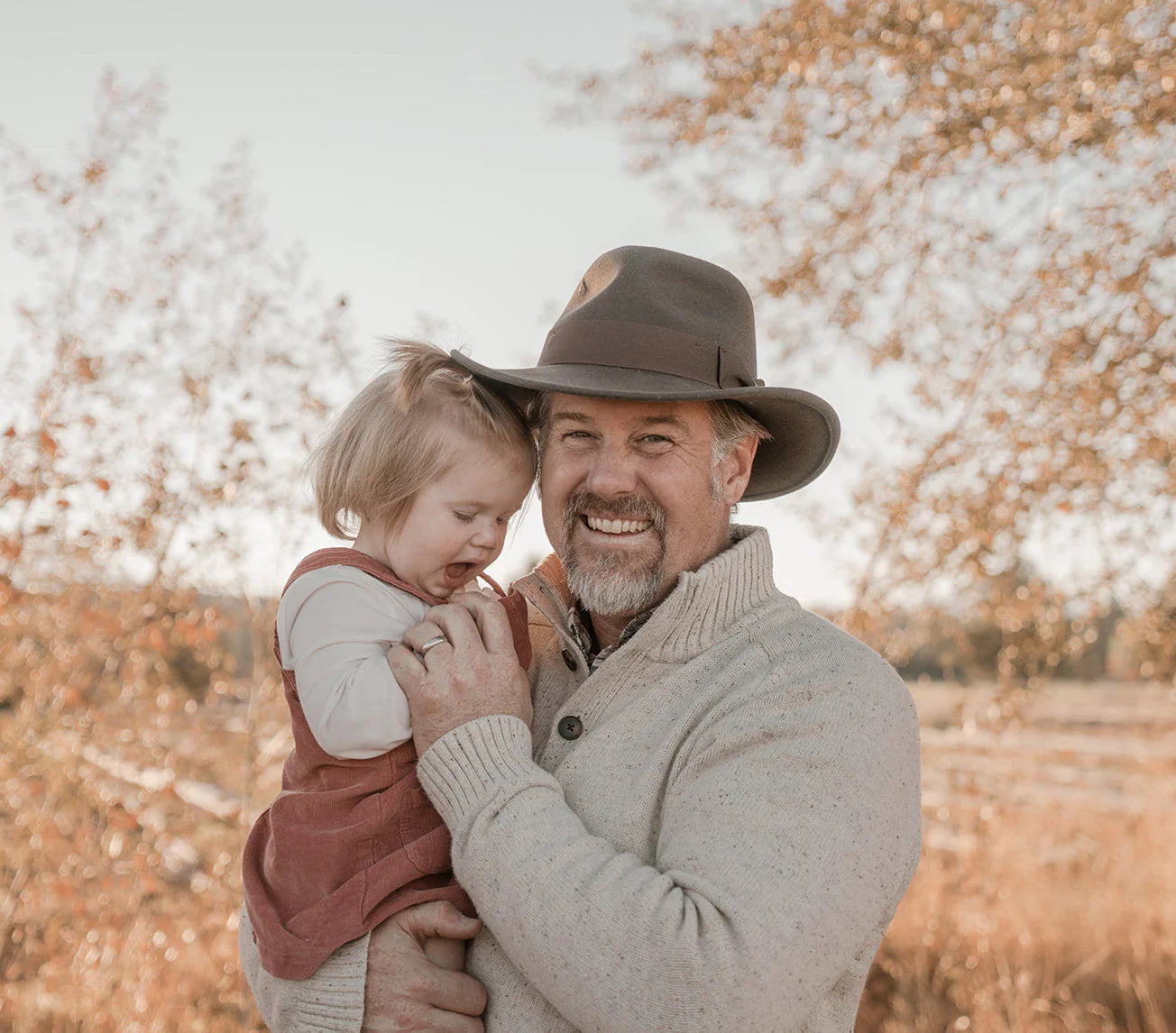 A smiling man in a wide-brimmed hat and beige sweater holds a laughing toddler wearing a red dress outdoors on a sunny, autumn day with golden trees in the background.