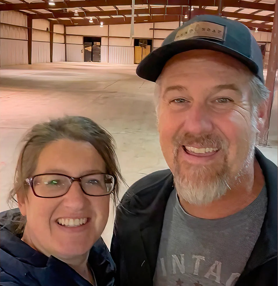 A smiling man and woman take a selfie inside a spacious, empty warehouse with high ceilings and exposed beams. The area is well-lit with fluorescent lights.