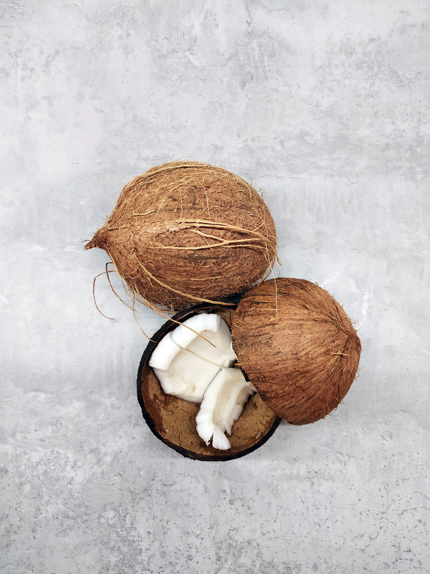 A whole coconut and a halved coconut with white coconut flesh inside are placed on a light gray textured surface.