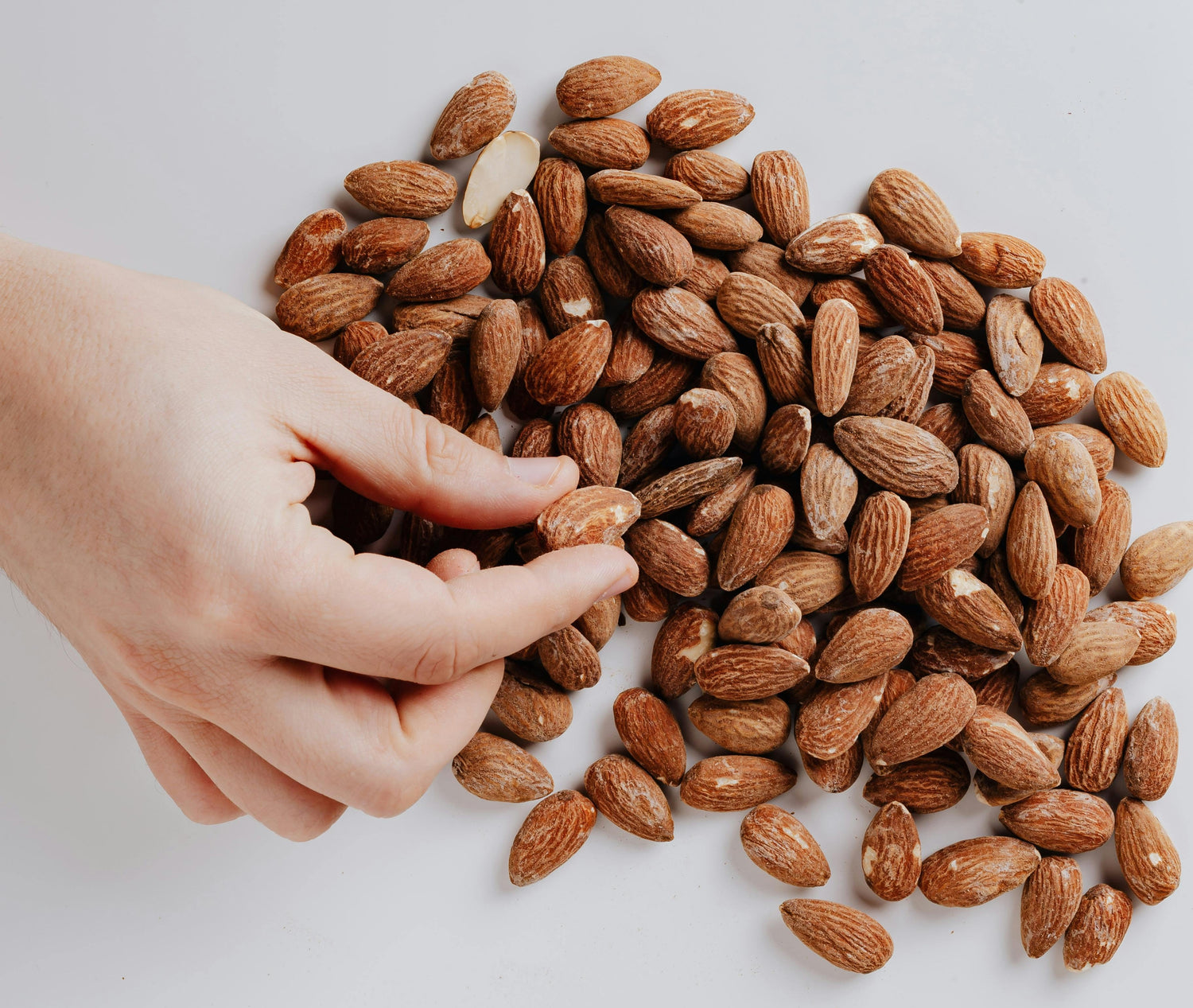 A hand picking up a raw almond from a pile of unshelled almonds on a white surface.
