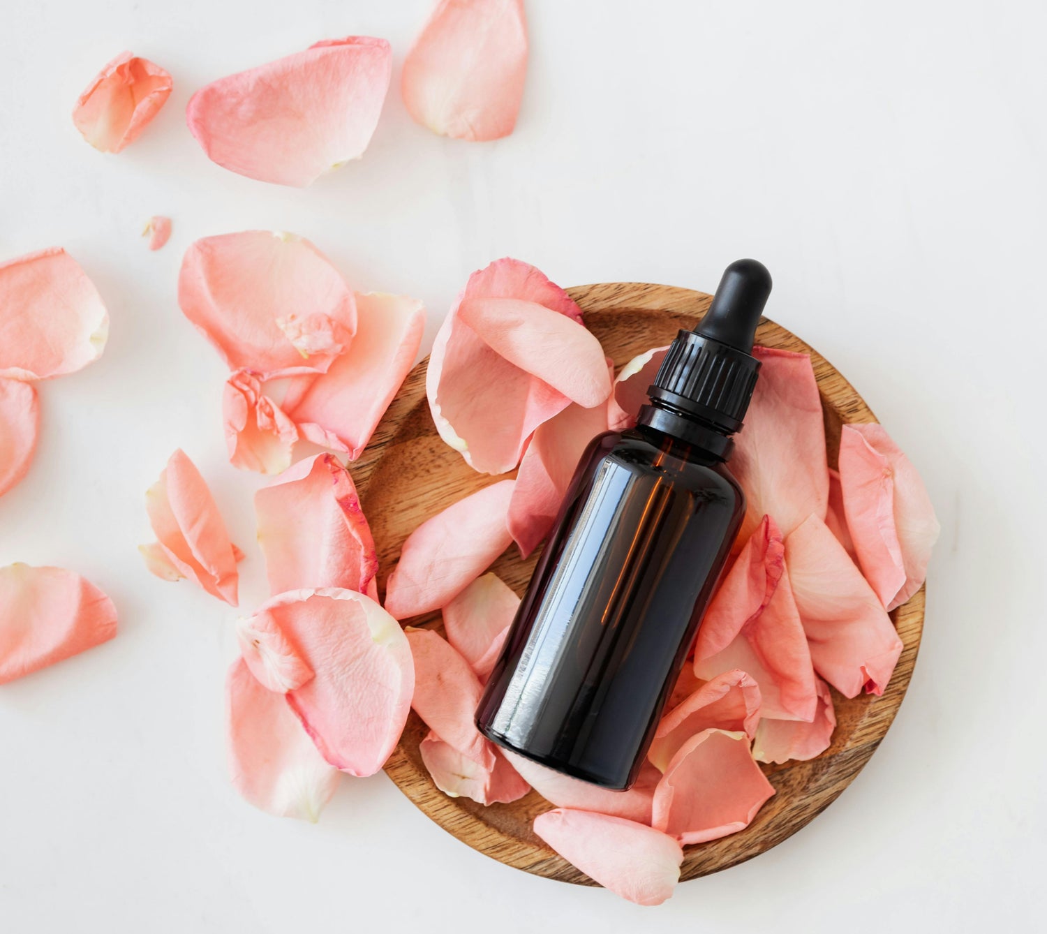 A dark glass dropper bottle sits on a wooden plate surrounded by scattered pink rose petals on a white surface.