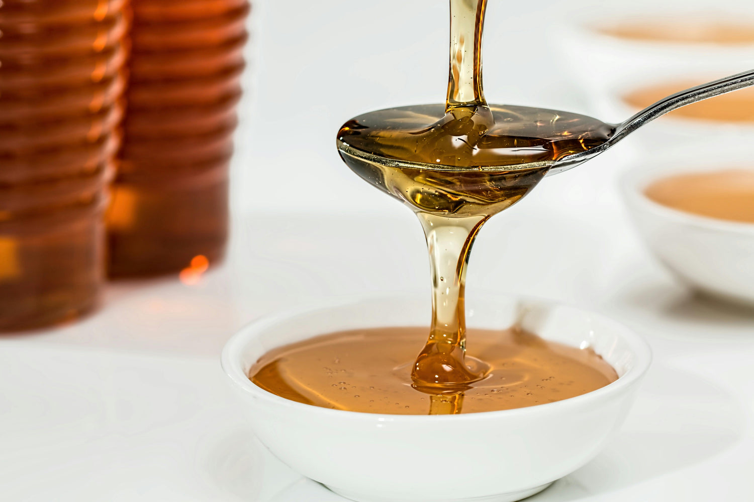 A spoon pours golden honey into a white bowl, with honey jars and more bowls in the blurred background on a white surface.