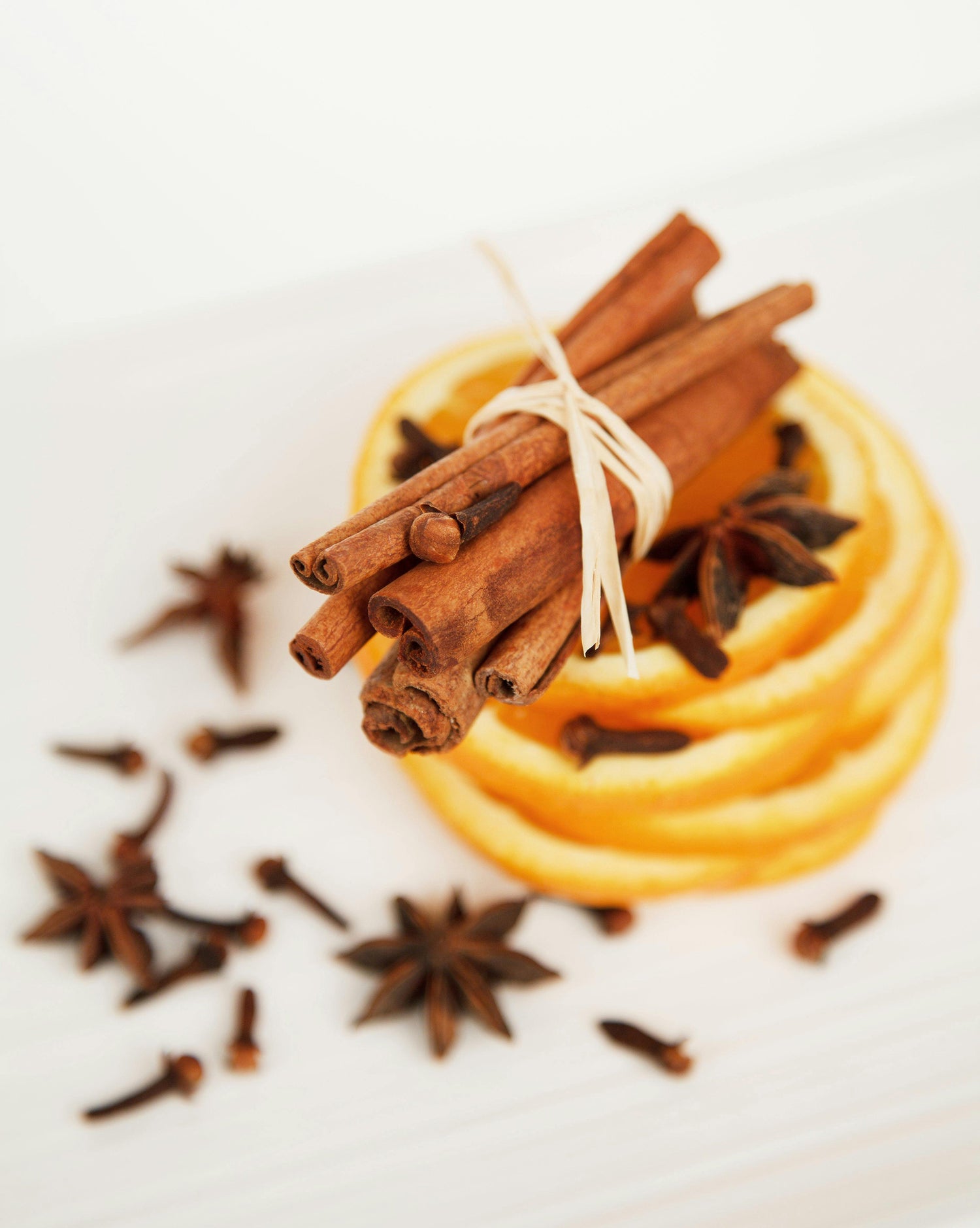 Slices of dried orange stacked with a bundle of cinnamon sticks tied together on top, surrounded by scattered star anise and cloves on a white surface.
