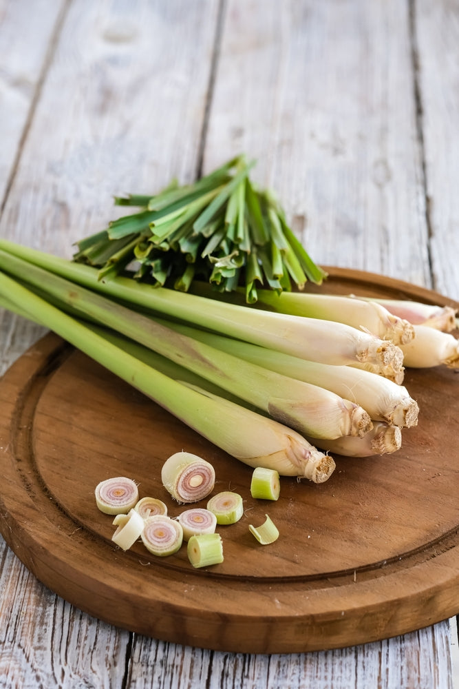 Fresh lemongrass stalks, sliced pieces, and leafy tops are arranged on a round wooden cutting board on a rustic wooden surface.