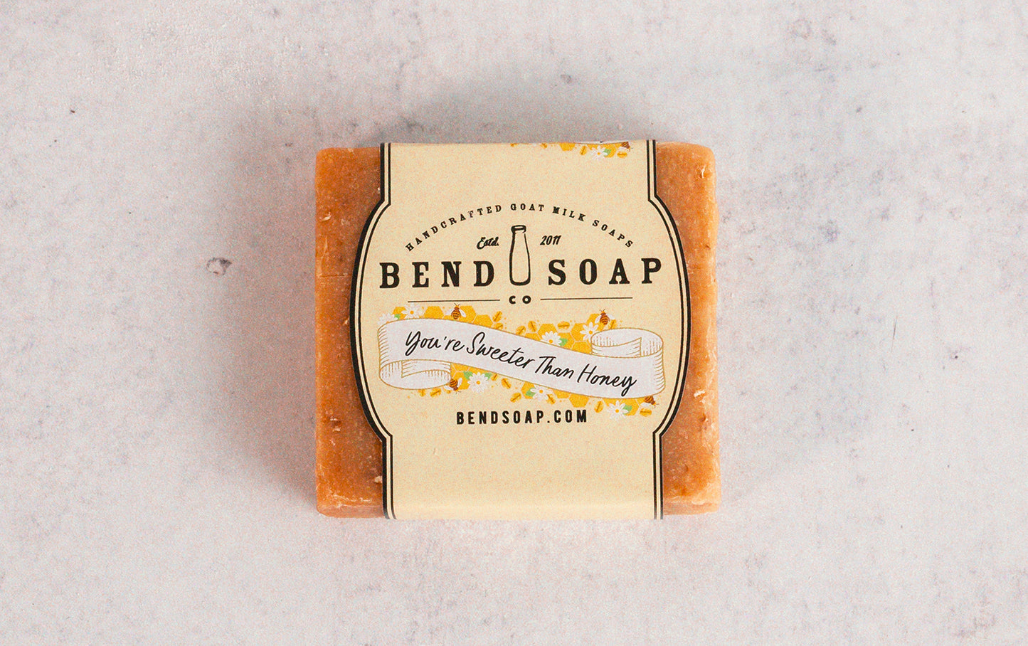 A Sweeter than Honey Gift Set from Bend Soap Co., featuring Honey Grapefruit goat milk soap with "You're Sweeter Than Honey" on the label and yellow floral accents, rests on a light marbled surface, suggesting a refreshing sugar scrub experience.