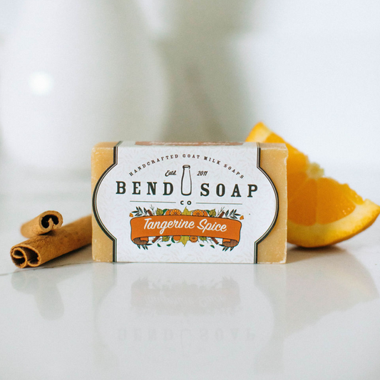 A bar of Tangerine Spice Goat Milk Soap rests on a white surface, with cinnamon sticks and an orange slice nearby, highlighting its natural tangerine oil for a refreshing skin boost.