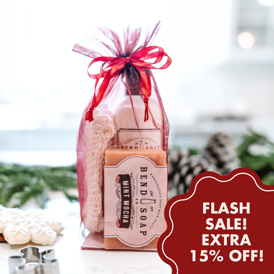 The Merry Little Gift Bag, featuring Goat Milk Soap and a washcloth in a sheer red bag, sits on a white surface. A red label says "FLASH SALE! EXTRA 15% OFF!" with pinecones and cookies softly blurred in the background.
