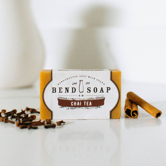 A 1 lb. portion of Chai Tea Goat Milk Soap Scrap rests on a white surface with its label showing, accompanied by two cinnamon sticks and cloves that highlight its sweet chai scent. The softly blurred background adds to the inviting scene.