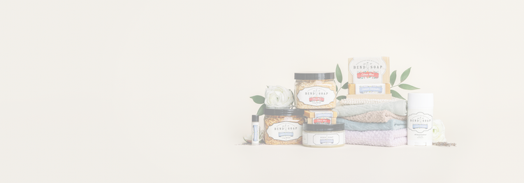 A collection of body care products, including jars, bottles, a bar of soap, and folded towels, arranged neatly with green leaves and a white flower on a light background.